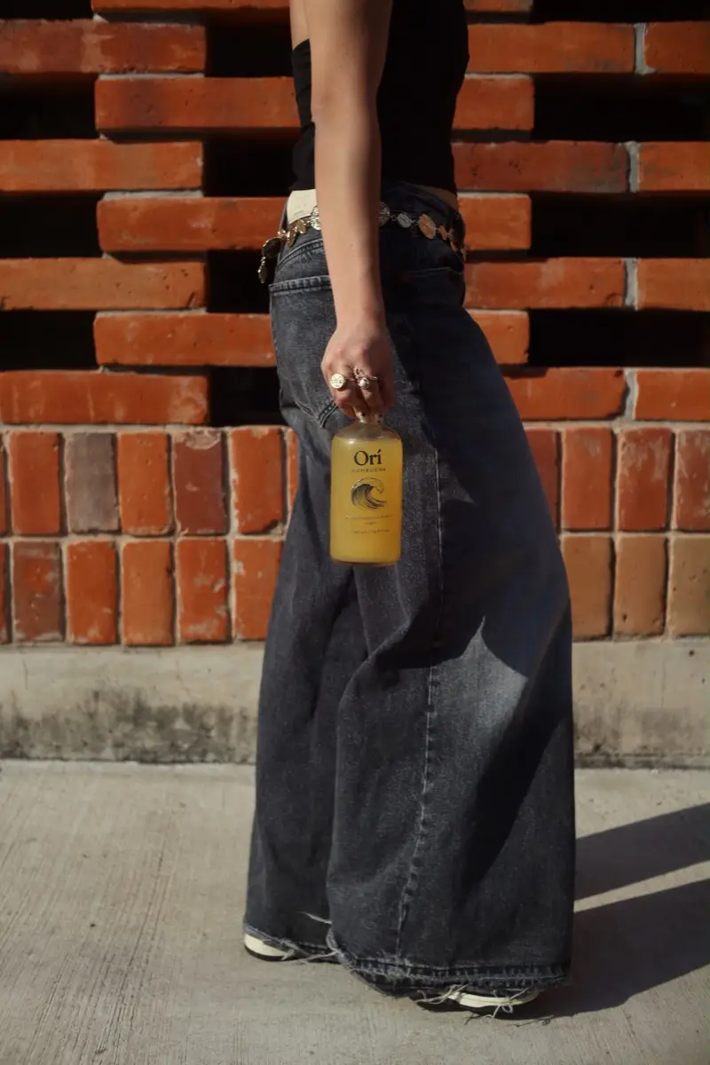 Botella de kombucha Orí a contraluz frente a una celosía de ladrillo, en la hora dorada